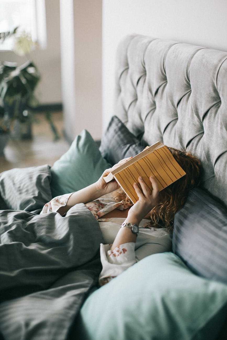 Woman in slept-in bed, holding a book over her face.